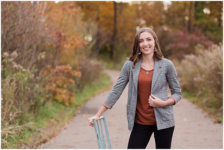 gorgeous waynedale high school female standing on path leaning on blue chair with lots of fall color behind her