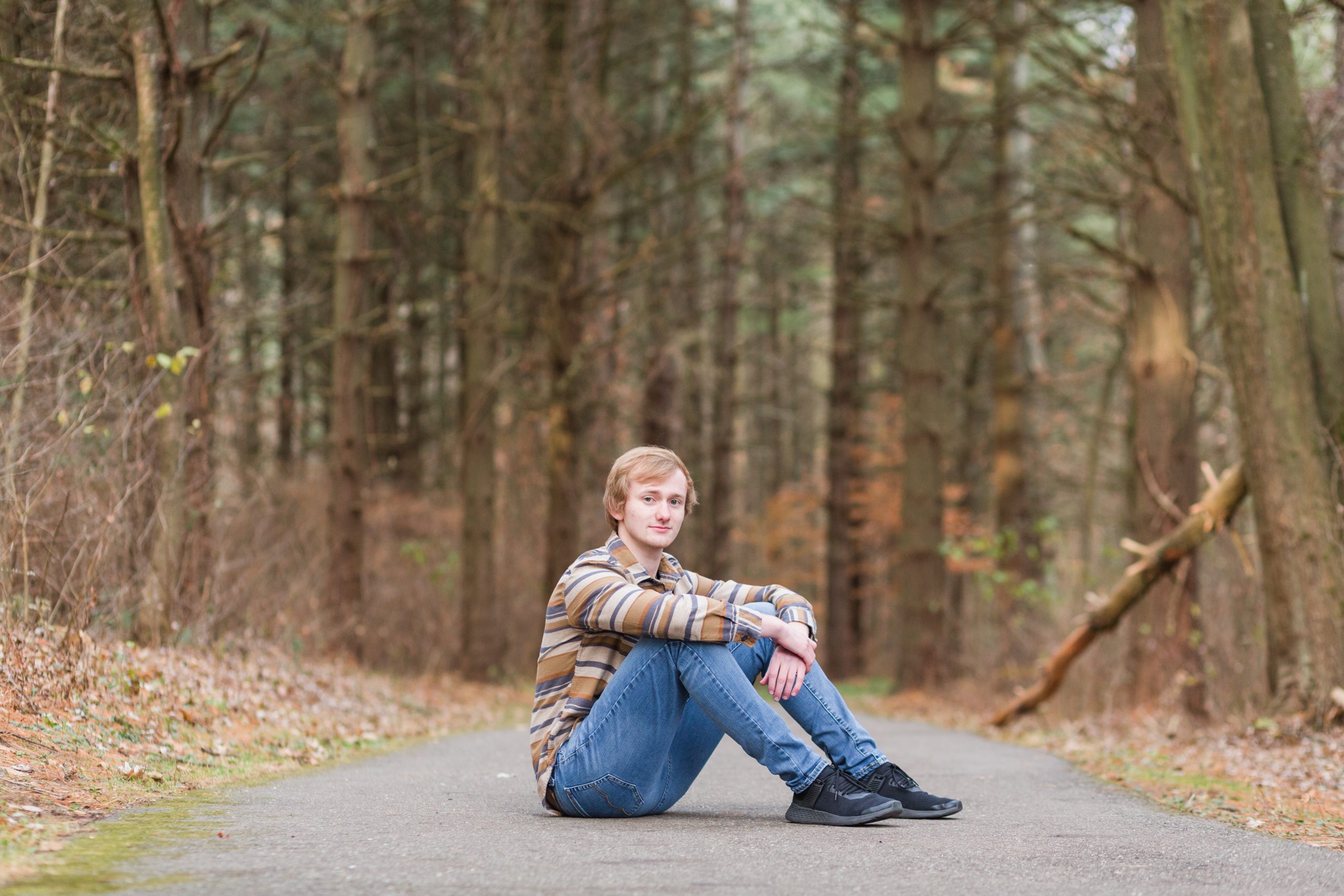 senior guy sitting on path in wooded area with leaves around and tall pines