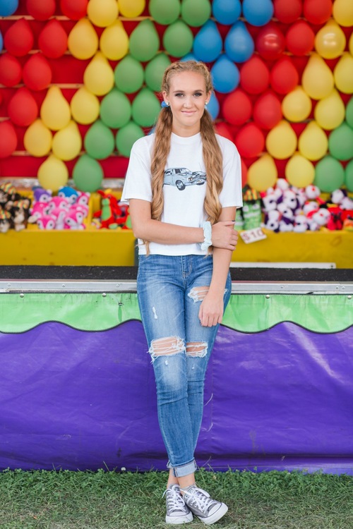 Senior girl smiling in front of the colorful balloon dart game booth at the Wayne County Fair, showcasing vibrant Wayne County Fair Senior Photos.
