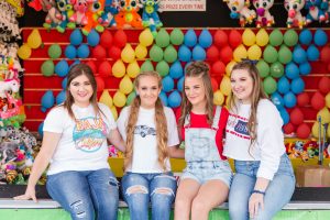 four senior high school girls sitting on the ledge of the ballon pop game at the fair, lots of beautiful colors behind the girls