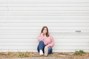 high school senior sitting on the ground in front of a white garage door she is wearing a pink sweater, she has one leg up and one down with a serious look on her face.
